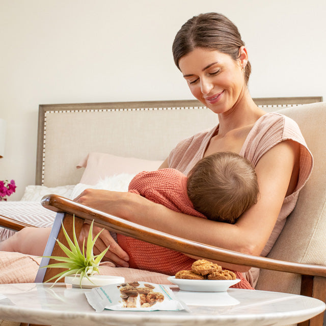 Woman breastfeeding a baby in a sling on a couch with snacks on a table in front